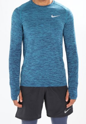 Mand iført en blå langærmet træningstrøje og sorte Nike-shorts, stående foran en ensfarvet baggrund.