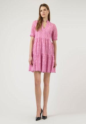 Nainen seisoo yllään vaaleanpunainen kirjailtu lyhyt mekko, jossa on puffy hihat, ja mustat korkokengät yksiväristä vaaleaa taustaa vasten.