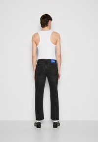 Jeans in denim nero con un taglio dritto, dotati di una patch con logo blu sul retro, indossati con una canotta bianca senza maniche e scarpe nere.