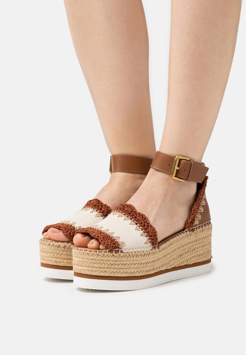 Pieds portant des sandales compensées crème et marron avec des détails tissés, des brides autour de la cheville et des semelles compensées en jute tressé.