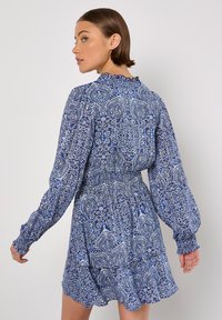 Langärmliges, blaues Kleid mit Muster, rüschigem Ausschnitt und gerafftem Taillenbereich. Verfügt über kunstvolle Blumen- und Paisleymuster.