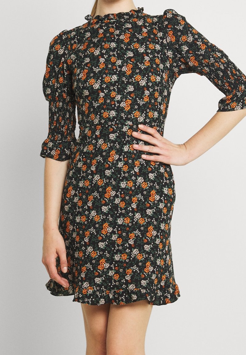 Femme portant une robe noire avec un motif floral orange et blanc, des manches bouffantes trois-quarts et un ourlet volanté, debout avec une main sur la hanche.
