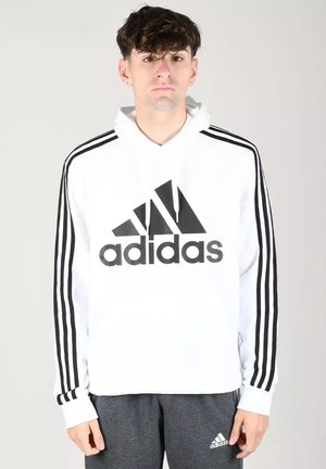 Giovane uomo che indossa una felpa con cappuccio bianca Adidas con strisce nere sulle maniche e pantaloni della tuta grigi Adidas, in piedi davanti a uno sfondo semplice.