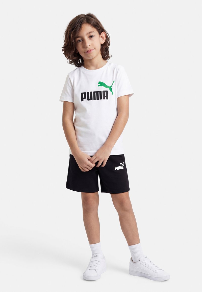 Παιδί που στέκεται φορώντας λευκό κοντομάνικο Puma, μαύρο σορτς Puma, λευκές κάλτσες και λευκά αθλητικά Puma, σε απλό φόντο.