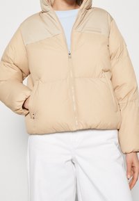 Beige prošívaná bunda se zipem, hladkým povrchem, kapucí a logem na prsou. Dvě přední kapsy.