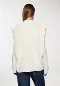 Maglione senza maniche a maglia color crema, con texture a coste, colletto alto e aperture per le braccia drop, abbinato a una camicia leggera a maniche lunghe e jeans blu.