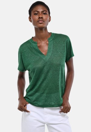 Femme portant un haut vert transparent à manches courtes avec un col en V échancré et un pantalon blanc, debout les mains dans les poches.