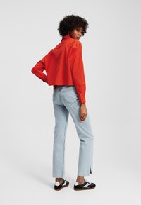 Rode cropped overhemd met lange mouwen, gecombineerd met lichtblauwe rechte jeans met een split aan de onderkant en zwart-witte sneakers.