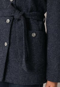 Tumši zils teksturēts mētelis ar austu rakstu. Ar piesaistāmu jostu, spīdīgām sudraba rozītes formas pogām un sānu kabatu.