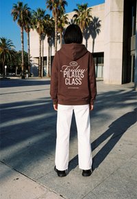 Personne aux cheveux noirs portant un sweat à capuche marron avec l'inscription « Friday Pilates Class » et un pantalon blanc, debout à l'extérieur près de palmiers et d'un bâtiment.
