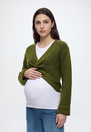 Pregnant woman with long brown hair wearing a green cardigan over a white shirt and blue jeans, holding her belly, standing against a white background.