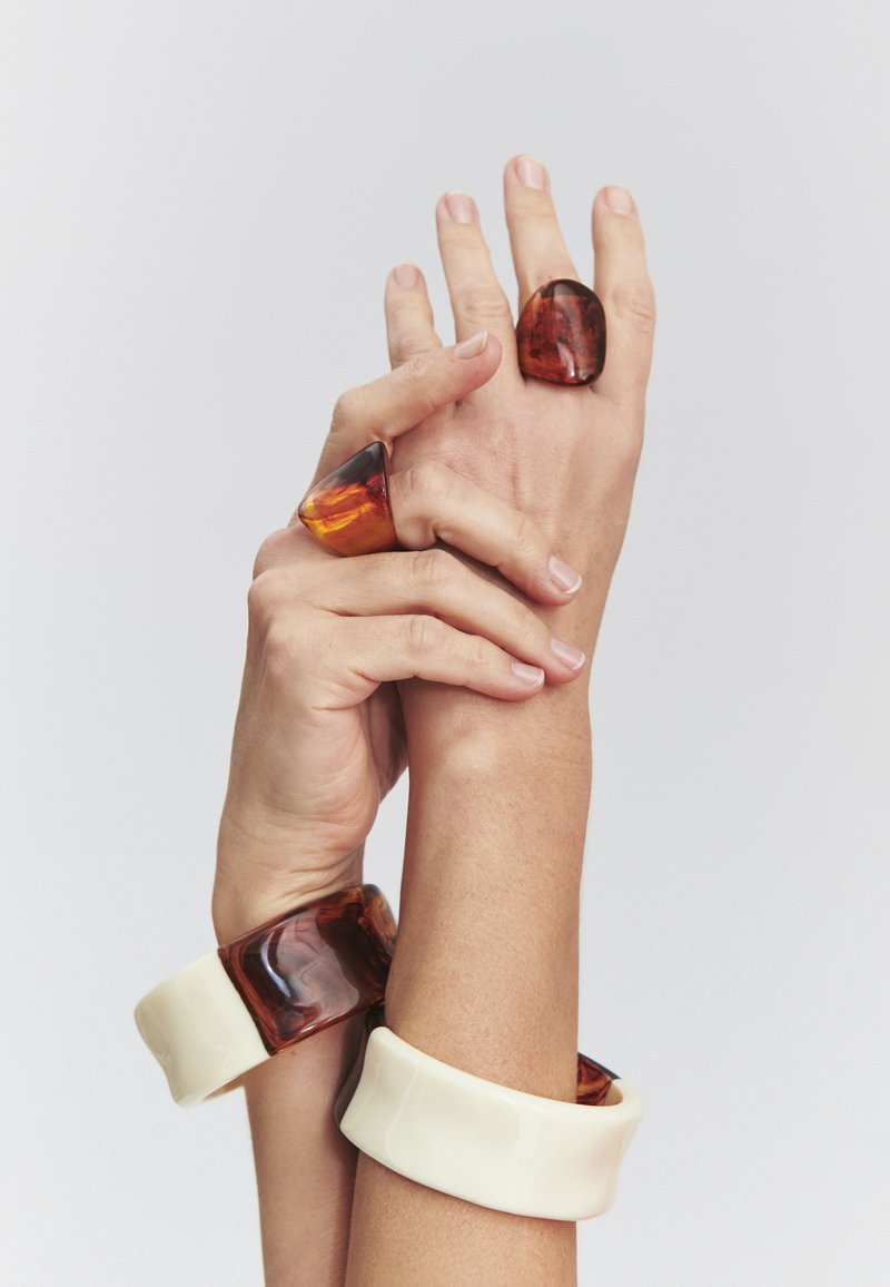 Deux mains portant des bagues en résine épaisses et des bracelets assortis dans des tons ambre et crème sur un fond uni.