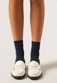 Białe skórzane mokasyny z czarnymi podeszwami, zestawione z ciemnoniebieskimi skarpetkami w kropki. Widoczna skóra od kostek do połowy łydki.