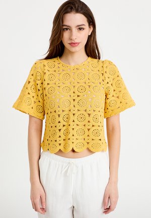 Joven mujer que lleva un top de crochet amarillo de manga corta con patrones circulares y borde ondulado, combinado con pantalones blancos con cordón.