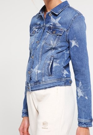 Chaqueta vaquera con estampados de estrellas blancas, llevada por una persona vestida con pantalones deportivos Reebok blancos, mostrada desde la mitad del torso hasta el cuello y la muñeca.