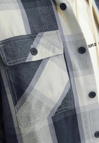 Primo piano di una tasca di camicia a quadri grigi e bianchi con bottone nero e bottoni neri visibili lungo il fronte della camicia.