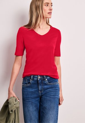 Kvinde iført rød kortærmet V-neck top og blå jeans, der holder en grøn jakke i venstre hånd og kigger mod højre.