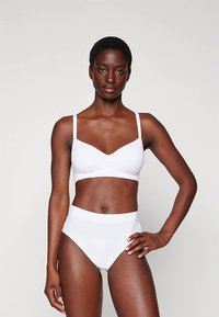 Kobieta z krótkimi włosami, ubrana w biały stanik i majtki z wysokim stanem, stoi z jedną ręką na biodrze na jednorodnym tle.