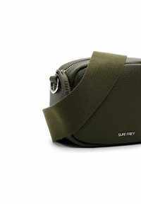 Borsa a tracolla in pelle verde texturizzata con una spessa cintura in tessuto, chiusura con zip e dettaglio del logo impresso. Forma rettangolare compatta.
