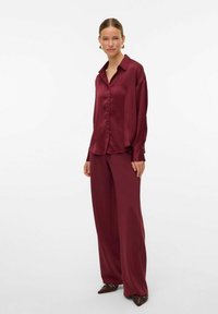Blusa de satén burdeos con cuello clásico, cierre de botones en la parte delantera y mangas largas, combinada con pantalones de pierna ancha a juego y zapatos de punta.
