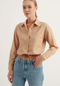 Camicia corta color beige con colletto a punta, taschino sul petto e dettaglio con cravatta in vita, abbinata a jeans azzurri chiari.