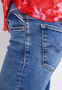 Nærbillede af blå denimjeans med Replay-mærkat på lommen, båret sammen med en rød bluse med blomsterprint.