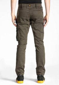 Pantalon cargo vert olive avec plusieurs poches latérales, design ajusté et détails de couture visibles. Porté avec des chaussures noires à semelles jaunes.