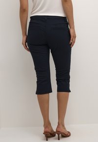 Námořnické capri kalhoty s hladkou texturou a mírným elastickým materiálem, které mají dvě malé zadní kapsy a zakončené lemy nad kotníky.