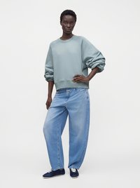 Personne debout, main sur la hanche, portant un sweat bleu clair oversize, un jean large bleu clair et des baskets bleu foncé sur fond uni.