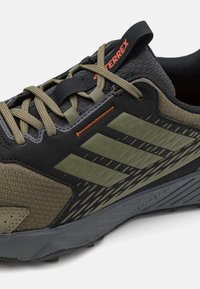 Zwarte en olijfgroene sportschoenen met een gestructureerde stof, rubberen zolen en drie horizontale strepen. Heeft een oranje accent op het label.