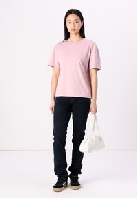 Jasnoróżowa bawełniana koszulka z kieszonką na piersi, zestawiona z ciemnoniebieskimi dżinsami i czarnymi trampkami z teksturowaną podeszwą, trzymająca białą torebkę.