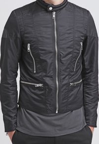 Svart vadderad jacka med hög krage, dragkedjefront och flera fickor med dragkedja. Slät textur med en strukturerad design.