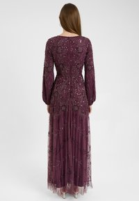Lungo abito da sera bordeaux con maniche lunghe, impreziosito da ricami floreali in perline e uno strato trasparente. Presenta una chiusura con zip sul retro.