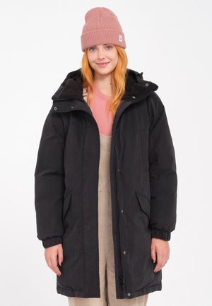Jeune femme aux longs cheveux roux porte un bonnet rose en tricot, un manteau matelassé noir avec capuche, une chemise rose et une salopette en velours côtelé beige sur fond blanc.