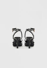 Melni augstpapēžu sandales ar kristālu rotājumiem, atklāta aizmugure un potīšu siksnas. Gluda tekstūra un slaidi papēži ērtai augstuma pievienošanai.