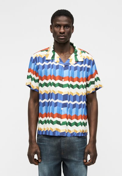 Άνδρας που φοράει κοντομάνικο πουκάμισο με πολύχρωμο γεωμετρικό ζιγκ ζαγκ μοτίβο και μπλε τζιν, στέκεται μπροστά σε απλό φόντο.
