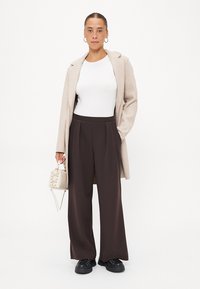 Beige kappa, vit åtsittande topp, mörkbruna vida byxor, svarta skor, håller en liten ljus väska. Släta texturer, skräddarsydd design och rena linjer.