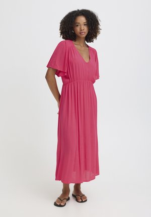 Kobieta stojąca w zwiewnej, różowej sukience midi z dekoltem w serek, z falbanowymi rękawami i czarnymi klapkami, na białym tle.