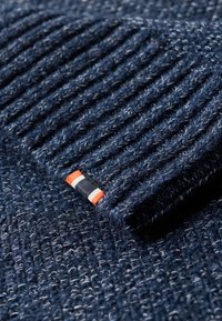 Gebreide trui in donkerblauw met een gestructureerd, ribbelpatroon. Voorzien van een klein label met rode, witte en blauwe strepen.