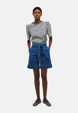 Femme portant un chemisier à manches bouffantes fleuri, un short en jean taille haute avec ceinture, et des ballerines lacées à motifs, debout les mains dans les poches.