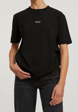 Personne portant un t-shirt noir à manches courtes avec le texte blanc « made to make a difference » centré, associé à un jean foncé.