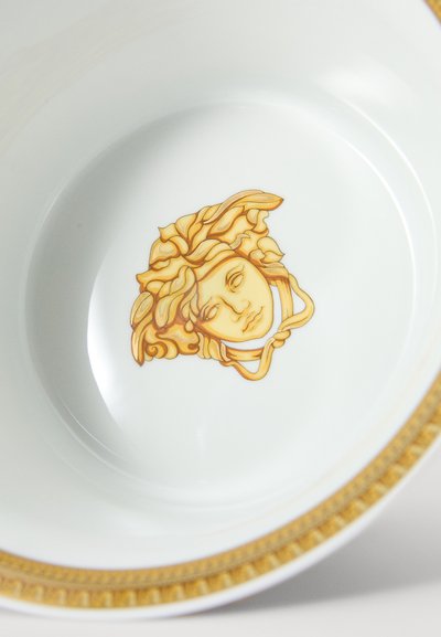 Prato de cerâmica branca com borda decorativa dourada e um emblema dourado da cabeça de Medusa no centro.