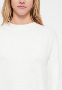 Balta džemperis ar brīvu piegriezumu, apaļu kakla izgriezumu un mīkstu tekstūru. Iekļauj nedaudz kritusī plecu daļu un bezšuvju dizainu.