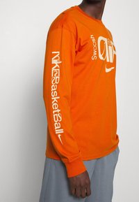 Long-sleeve orange T-shirt made of cotton, featuring white Nike branding and graphics on the front and sleeve, with a relaxed fit.
