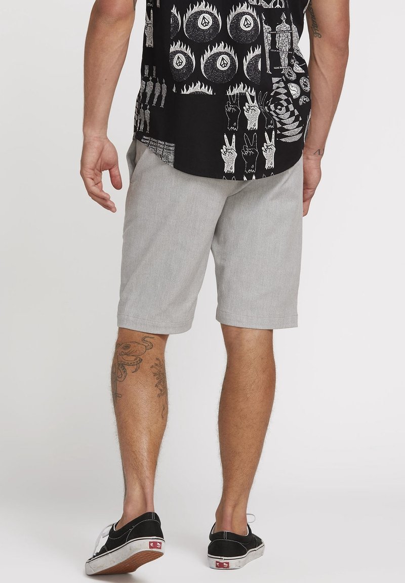 Homme portant un short gris clair, une chemise noire imprimée de signes de paix et de flammes, des baskets noires basses et des tatouages sur les jambes, debout de dos à la caméra.