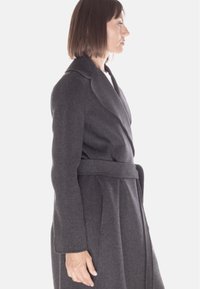 Cappotto trench in lana grigia con vita slacciata, ampi risvolti, forma su misura e tasche laterali. Texture liscia e maniche lunghe.