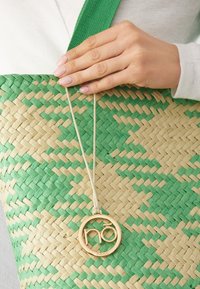Geflochtene Strohtasche mit einem grünen und beigen Muster, ausgestattet mit einem goldfarbenen Logo-Anhänger, der an einer beigen Schnur an der Seite befestigt ist.