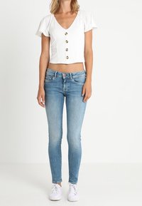 Top corto blanco de manga corta con escote en V y detalles de botones, combinado con jeans ajustados azul claro y zapatillas blancas.