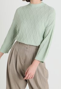 Ljusgrön stickad tröja med diamantmönster, hög rund halsringning och löst sittande ärmar, ihop med beige randiga byxor.