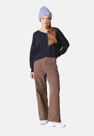 Femme portant un bonnet bleu clair en maille, un haut noir à manches longues, un pantalon large en velours côtelé marron et des baskets blanches, debout avec une main sur la hanche.